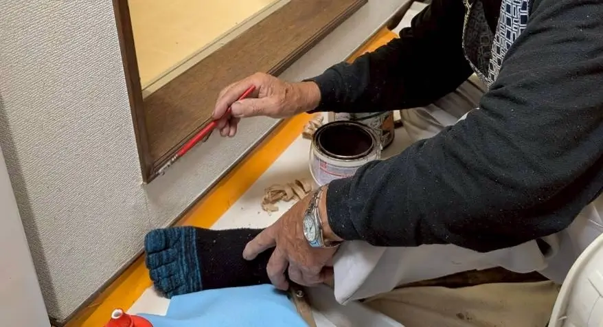 浴室出入口の木製ドア枠を補修後、刷毛で塗装仕上げを行う職人の様子。木部の色合わせや質感を整える丁寧なリフォーム工程。